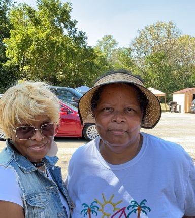 Two women smiling outdoors on a sunny day, standing beside a parked car.