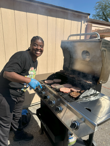 Person grilling hamburgers on a gas grill outdoors, smiling with blue gloves on.