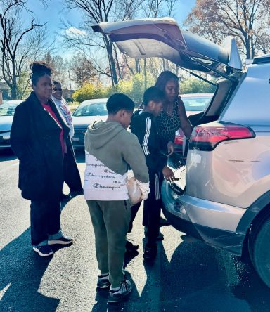 A family gathers around the open trunk of a car, engaging in conversation and activity.