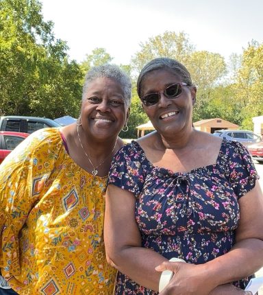 Two smiling women pose together outdoors, wearing colorful tops against a sunny background.