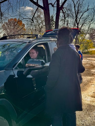 A person in a car interacts with a person standing outside. Trees are visible in the background.