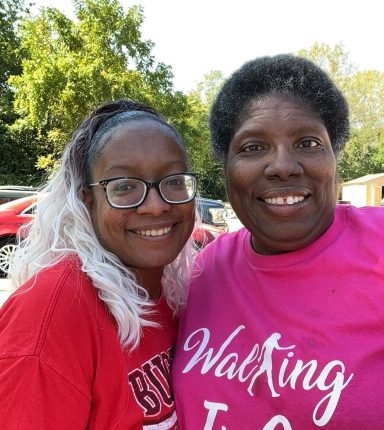 Two women smiling together outdoors, one wearing a pink shirt and the other in red.