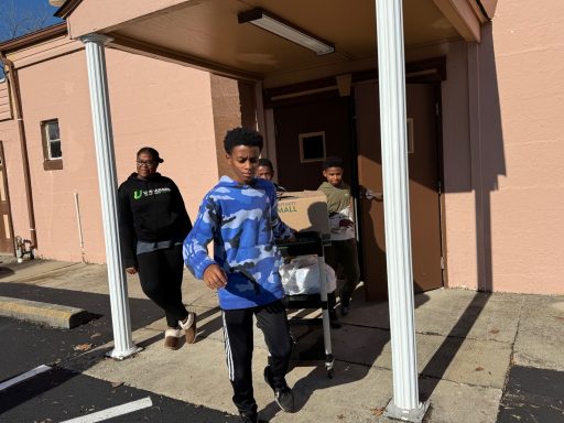 Three children leave a building, one carrying a large bag, on a sunny day.