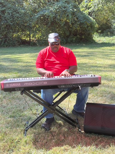 A man in a red shirt playing a keyboard outdoors on a sunny day.