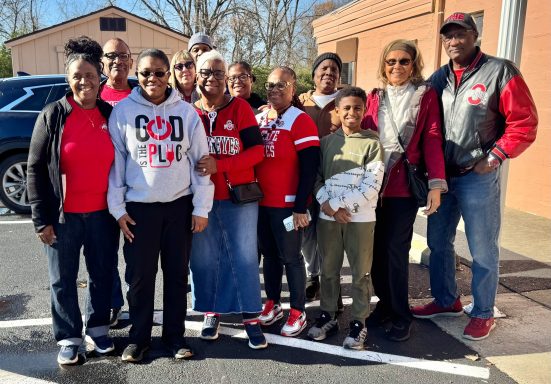 A diverse group of people smiling together outdoors, wearing casual clothing in red and white.