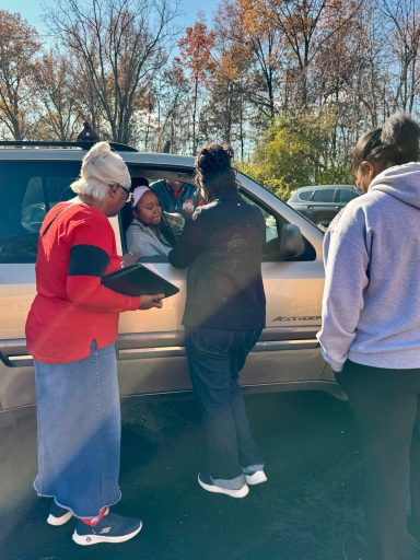Four people interact near a parked vehicle in a sunlit outdoor setting.