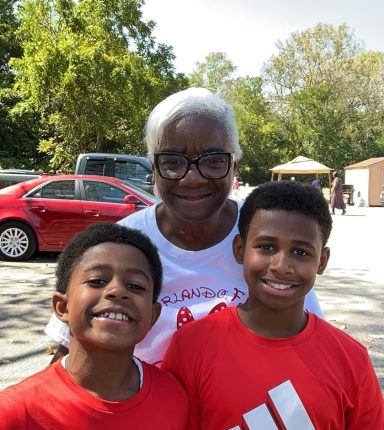 Two boys smiling with a woman in a white shirt, outdoors with trees in the background.