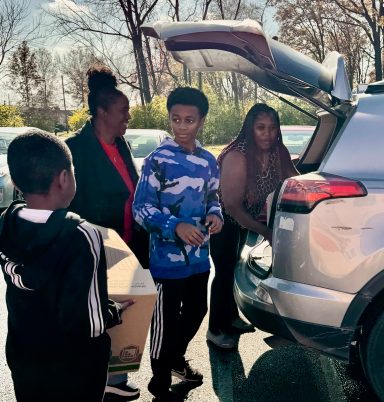 A woman and two children load boxes into the back of a car in a parking lot.