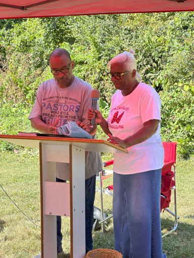 Two people speaking at an outdoor event, one holding a microphone and the other reading from notes.