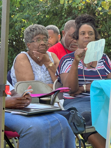 A group of people sitting outdoors, engaged in conversation and holding papers.