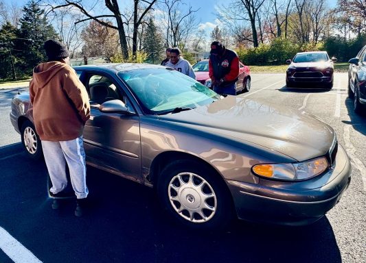 Two people converse near a gray car parked in a lot on a sunny day.