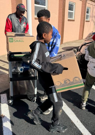 Children carrying boxes during a community event with an adult supervising.