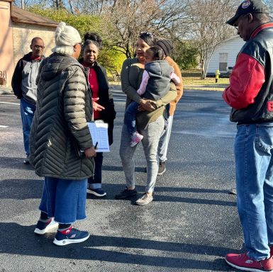 A group of people talking outdoors, some holding a child, in a sunny neighborhood.
