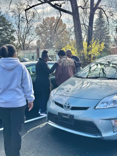 Four people interact near parked cars on a tree-lined street in daylight.