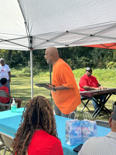 A man in an orange shirt stands at a table during an outdoor gathering.